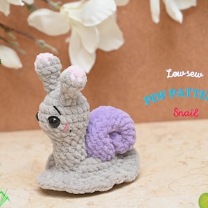 Op de afbeelding: Een gehaakt amigurumi-slak speelgoed met een paarse schelp en een grijs lichaam. De slak heeft een schattig gezicht met zwarte ogen en roze wangen. Het speelgoed is een PDF-patroon voor een low-sew project.