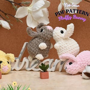 Może przedstawiać: Trzy pluszowe zabawki amigurumi w kształcie królika, wykonane na szydełku, w różnych kolorach: szarym, białym i różowym. Króliki siedzą na białym znaku z napisem "dream". Znak stoi na brązowej powierzchni z zielonymi roślinami i kulkami z waty.