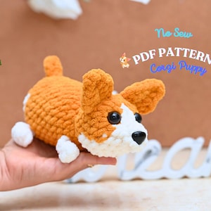 May include: A crocheted corgi puppy in shades of orange, white, and black. The plush toy has large ears, a small tail, and black button eyes. The image includes the text "No Sew PDF Pattern Corgi Puppy."