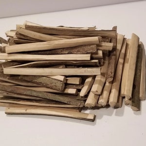 Peut inclure: Un tas de bâtonnets de bois brun clair, certains avec de l'écorce. Les bâtonnets sont de tailles et de formes variées, certains semblent coupés ou sculptés. Le bois a un aspect naturel et non traité.