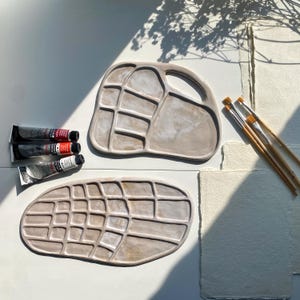 Puede incluir: Dos paletas de pintura de cerámica con pozos de diferentes tamaños, una con asa. Las paletas son de color marrón claro con un glaseado blanco. Tres tubos de pintura están sobre una superficie blanca junto a las paletas. Cuatro pinceles están sobre una superficie blanca junto a las paletas.