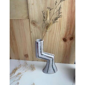 May include: A white ceramic vase with a geometric design and black stripes. The vase is shaped like a leg and has a wide base. It is filled with dried flowers.