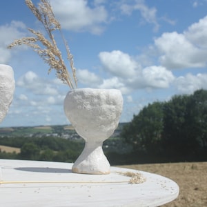May include: Four white ceramic vases with a textured surface, arranged on a white wooden table. The vases are in the shape of a goblet with a wide bowl and a narrow base. The vases are filled with dried grasses and are set against a backdrop of a blue sky with white clouds and a green field.