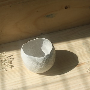 Può includere: Una ciotola in ceramica bianca con una superficie strutturata e una forma irregolare. La ciotola è posizionata su una superficie di legno, con fiori secchi e ombre. L'interno della ciotola è liscio, in contrasto con la texture ruvida dell'esterno.