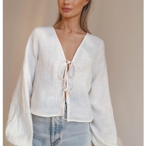 May include: White linen blouse with a V-neck and long sleeves. The blouse has a tie at the front.
