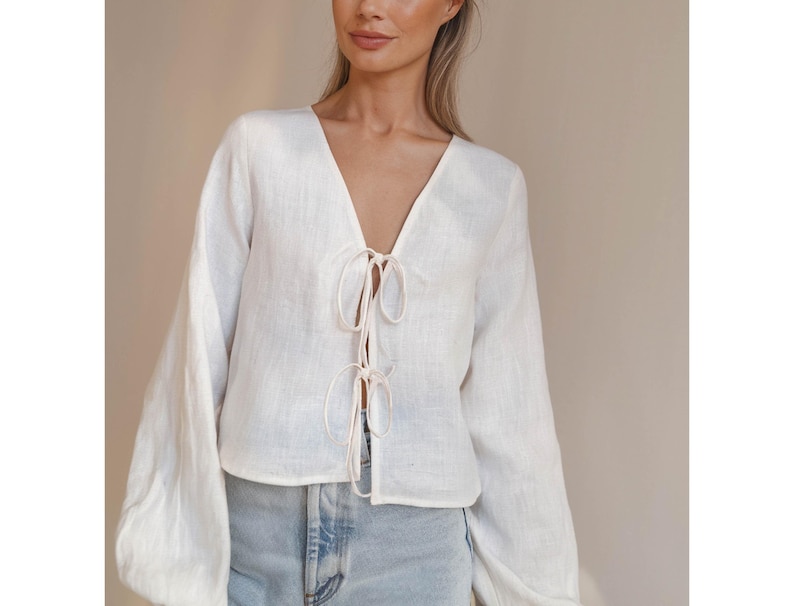 May include: White linen blouse with a V-neck and long sleeves. The blouse has a tie at the front.