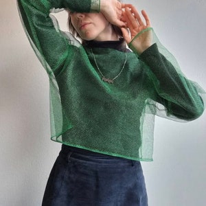 May include: A green mesh long-sleeved shirt with a high neckline. The shirt is made of a sparkly material and has a loose fit.