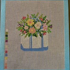 May include: A square needlepoint canvas featuring a blue basket filled with a colourful floral arrangement of yellow, pink, and green stitches. The canvas is bordered by light blue fabric tape and includes a vertical colour palette guide on the left.