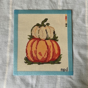 May include: A needlepoint canvas featuring a stack of two pumpkins, one orange and one cream-colored, surrounded by green leaves. The canvas has a light blue border, a small color palette guide in the upper right corner, and the letters npd in the corner.