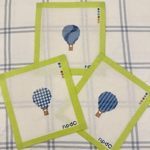 May include: Three square needlepoint canvases with lime green fabric borders. Each canvas features a different blue hot air balloon design with a brown basket. Two canvases include the text npdc in the bottom corner and color swatches.