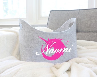 Peut inclure: Un panier de rangement en feutre gris avec une licorne rose et le nom "Naomi" imprimé dessus. Le panier est posé sur une couverture blanche et moelleuse.