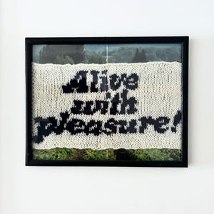 May include: Framed artwork with the words "Alive with pleasure!" in black on a cream knitted texture. The artwork is set against a landscape and framed in a black frame. The artwork is displayed on a white wall.