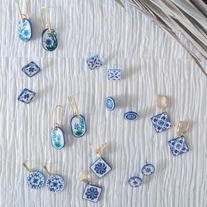 May include: A collection of blue and white earrings in various shapes, including oval, square, and round. Some earrings feature floral designs, while others have geometric patterns. Gold-colored earring hooks and posts are visible.