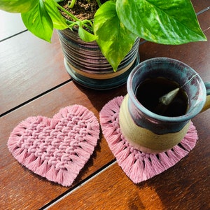 May include: Two pink heart-shaped crocheted coasters with fringe, one with a mug of tea on top. The mug is a dark blue and brown ceramic with a tea bag in it. The coasters are on a wooden surface.