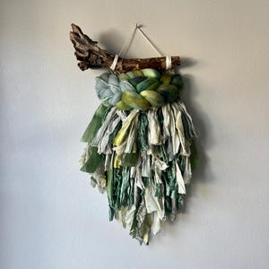 May include: A green, white and blue woven wall hanging with a driftwood branch at the top. The hanging has a fringe of fabric at the bottom.