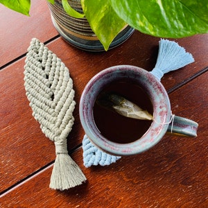 May include: A close-up of a cup of tea with a tea bag in it, sitting on a light blue and grey leaf-shaped coaster. The cup is on a wooden table with a green plant in the background.