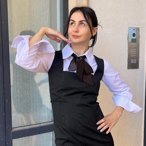 May include: A black pinstriped sleeveless dress with a square neckline and a side slit, worn over a crisp white long-sleeved button-down shirt. The outfit is accented with a dark brown satin necktie tied in a bow at the collar of the white shirt.