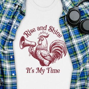 Pode incluir: Camiseta branca com uma ilustração de um galo vermelho segurando um megafone. O texto "Rise and Shine It's My Time" está impresso na camiseta.