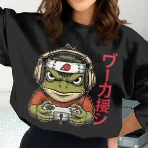 Puede incluir: Sudadera negra con un dibujo de una rana con auriculares y una diadema, sosteniendo un mando de videojuegos. La rana es verde con una expresión enfadada. Texto japonés impreso en el lado derecho de la sudadera.