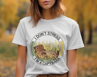 Puede incluir: Camiseta blanca con un gráfico de una capibara de dibujos animados tumbada en un campo de flores amarillas. El gráfico está rodeado de un círculo con el texto "No me estreso, soy una capibara".