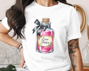 May include: White t-shirt featuring a watercolor illustration of a glass bottle filled with pink liquid, labeled "Love Potion." The bottle has a cork stopper and is adorned with a black ribbon. The design is centered on the shirt.