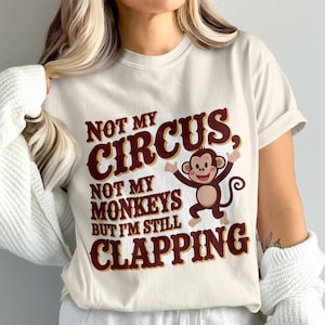 May include: Cream-colored t-shirt with the phrase "NOT MY CIRCUS, NOT MY MONKEYS BUT I'M STILL CLAPPING" in maroon lettering. A cartoon monkey with outstretched arms is featured in the design.
