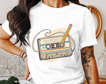 May include: White t-shirt featuring a retro cassette tape design with a pencil inserted. The cassette has a striped design in shades of red, yellow, blue, and green. The pencil is yellow with a pink eraser. The design is set against a light beige circle.