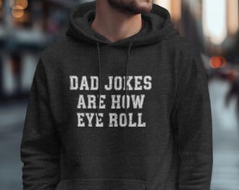 May include: Dark gray hoodie with the text "DAD JOKES ARE HOW EYE ROLL" in white, distressed lettering. The hoodie has a drawstring and a front pocket. The background is blurred, suggesting an outdoor setting.