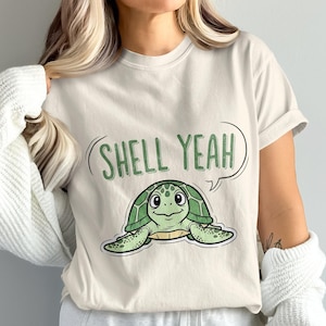 Könnte beinhalten: Cremefarbenes T-Shirt mit einer Cartoon-Schildkröte mit grünem Panzer und dem Text "SHELL YEAH" in einer Sprechblase. Das Shirt hat einen lässigen Stil und ist für den Alltag geeignet.