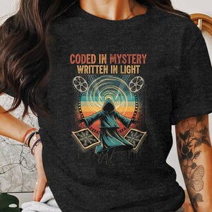May include: Dark gray t-shirt featuring a graphic with the text "CODED IN MYSTERY WRITTEN IN LIGHT." The design includes a robed figure, film reels, and geometric shapes in a retro color palette. A unique graphic tee.