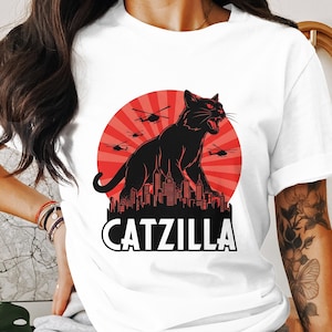 May include: White t-shirt featuring a black illustration of a cat in a city, with the text "CATZILLA". The cat is depicted roaring with red eyes, with helicopters in the background against a red sunburst design.