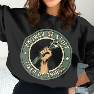 Pode incluir: Camisola preta com um design circular verde e creme. O design apresenta o texto "KNOWer OF STUFF FIXER OF THINGS" e uma mão segurando uma chave inglesa.