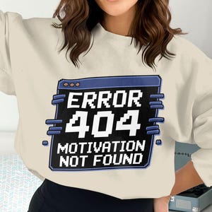 May include: A cream-coloured sweatshirt with a pixelated graphic displaying "ERROR 404 MOTIVATION NOT FOUND" in white and blue. The design is set within a blue-bordered window frame, giving a retro computer error message aesthetic.