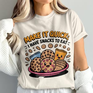 Puede incluir: Camiseta color crema con un gráfico colorido de galletas y una bolsa de snacks en un plato rosa. El texto "MAKE IT QUICK; I HAVE SNACKS TO EAT" está en naranja y negro. La camiseta es de uso diario.