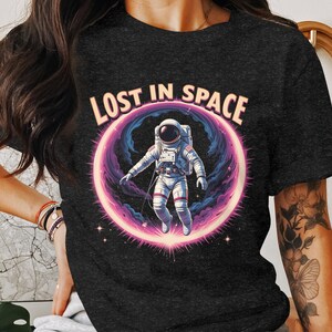 May include: Dark gray t-shirt featuring a colorful graphic of an astronaut floating in a pink and purple space portal. The text "LOST IN SPACE" arches above the astronaut in a light orange color.