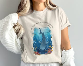 May include: Cream-colored t-shirt featuring a detailed illustration of a whale shark swimming in the ocean. The design includes colorful coral, fish, and bubbles. Text above the image reads, "When you go through deep waters, I will be with you - Isaiah 43:2."