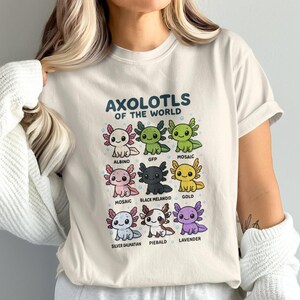 May include: Cream-colored t-shirt featuring a colorful graphic of various axolotl types, including Albino, GFP, Mosaic, Black Melanoid, and Gold. The text "AXOLOTLS OF THE WORLD" is printed above the axolotl illustrations. The shirt is made of a soft material.