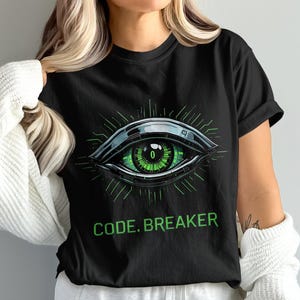 Peut inclure: T-shirt noir avec un motif d'œil stylisé en vert et argent. L'œil a un aspect numérique avec le chiffre "0" au centre. Le texte "CODE. BREAKER" est imprimé en vert en dessous.