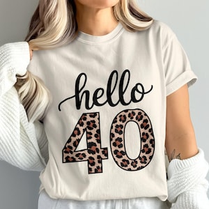 May include: Cream-colored t-shirt with the words "hello 40" in black script. The numbers "40" are in a leopard print pattern. The shirt is a casual style.