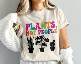 Könnte beinhalten: Cremefarbenes T-Shirt mit dem Text "PLANTS, NOT PEOPLE" in farbenfrohen Buchstaben im Retro-Stil. Darunter befinden sich drei schwarze Topfpflanzen: eine hohe, schlanke Pflanze, eine Monstera und ein Kaktus. Ein lässiges Design zum Thema Pflanzen.
