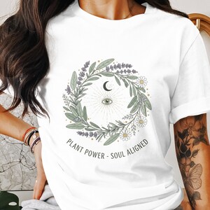 Peut inclure: T-shirt blanc avec un motif de couronne botanique, comprenant un croissant de lune, un œil et le texte "PLANT POWER - SOUL ALIGNED". La couronne comprend de la lavande, des marguerites et des feuilles vertes.