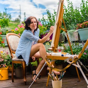 May include: A woman sits on a chair in a garden setting, painting on a canvas easel. She is wearing a light blue shirt and has her hair pulled back. The easel is set up on a wooden stand with a variety of paintbrushes in a cup.