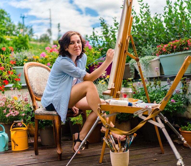 May include: A woman sits on a chair in a garden setting, painting on a canvas easel. She is wearing a light blue shirt and has her hair pulled back. The easel is set up on a wooden stand with a variety of paintbrushes in a cup.