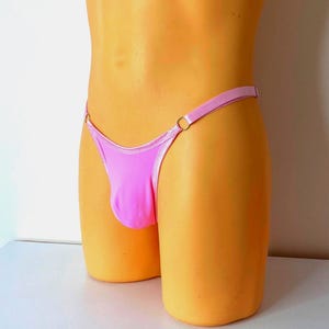 Puede incluir: Un tanga rosa con una fina cintura y una pequeña bolsa. El tanga presenta un detalle de anillo en cada lado. El traje de baño se exhibe en un maniquí.
