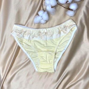 Könnte beinhalten: Hellgelbe Unterwäsche mit weißer Bordüre und floraler Stickerei am oberen Rand. Die zarte Dessous liegen auf einem Satinhintergrund mit Akzenten von Baumwollzweigen.