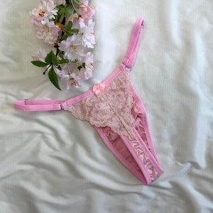 Op de afbeelding: Een lichtroze string met een delicate kanten bloemenoverlay op het voorpand, dunne verstelbare bandjes en een klein bijpassend satijnen strikje in het midden van de tailleband. Het kledingstuk is gemaakt van een zachte, gladde stof met een subtiele glans.