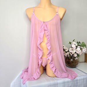 Può includere: Un completo intimo rosa chiaro e trasparente su un manichino. Il set include un abito fluido con apertura frontale e bordi a balze e un perizoma abbinato. L'abito ha spalline delicate e piccoli dettagli floreali.