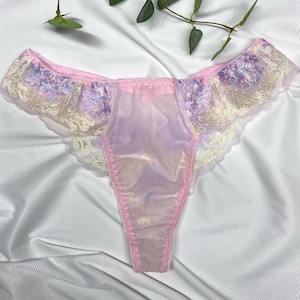 Könnte beinhalten: Ein zarter String-Slip in Rosa- und Lavendeltönen. Die Ränder sind mit floraler Spitze in Creme- und Pastellfarben verziert. Der transparente Stoff zeigt einen weichen, irisierenden Glanz.