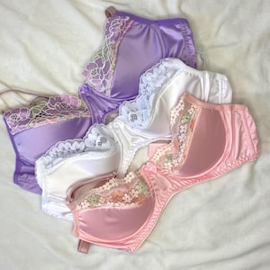 May include: Four satin bras in shades of purple, white, and pink are arranged on a white surface. Each bra features lace detailing along the cups and straps. The bras have underwire support and adjustable straps.