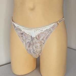 Può includere: Perizoma avorio con dettagli in pizzo floreale. Il pannello frontale è in materiale satinato e i bordi sono rifiniti con un delicato pizzo smerlato. Le spalline sono sottili e metalliche.